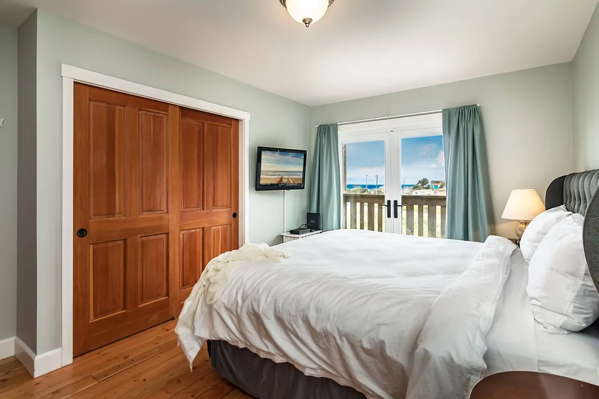 A serene bedroom featuring a comfortable bed with white linens, a nightstand with a lamp, wooden closet doors, a flat-screen TV, and large windows that offer a partial view of the sea. The soft, natural lighting creates a calm and inviting atmosphere perfect for relaxation or private downtime.