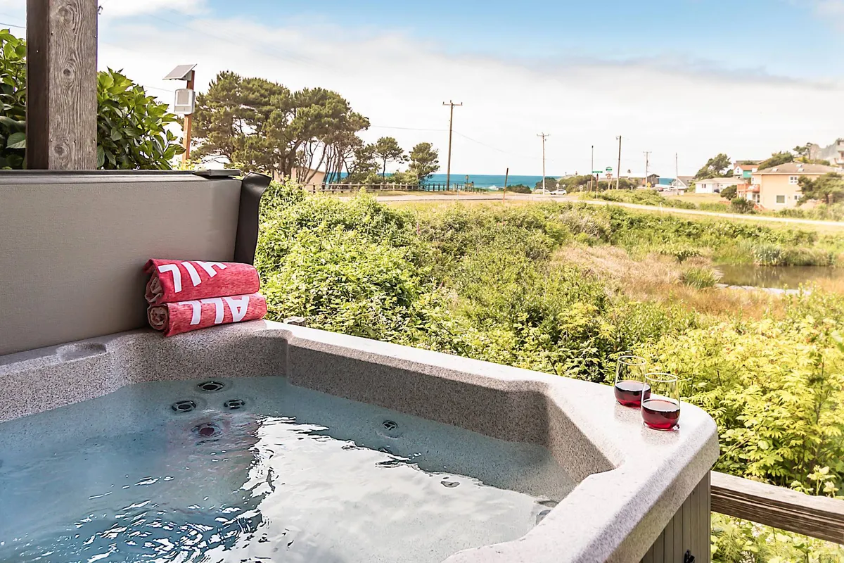 A tranquil exterior setting featuring a bubbling hot tub with fresh towels nearby, set against a scenic backdrop of wetlands and distant ocean views. The patio area invites relaxation, socializing, and soaking up the natural beauty just steps outside the entertainment room.