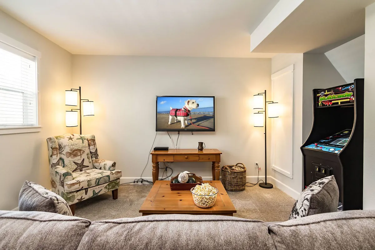 A cozy entertainment space featuring a large television displaying a playful image, a wooden coffee table with popcorn and snacks, an arcade machine tucked into the corner, and a stylish decorative chair. The room is set up for movie nights, game tournaments, and casual lounging, capturing a fun and welcoming vibe.