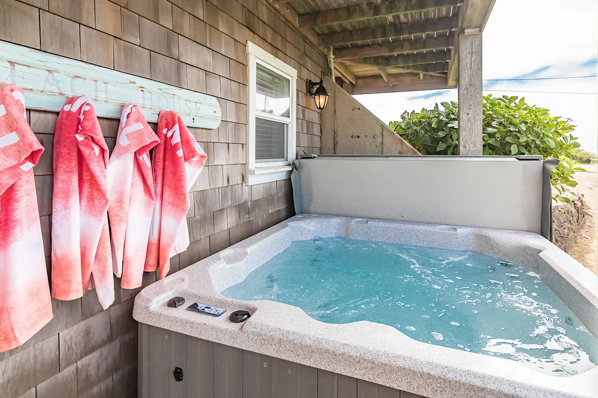 An exterior view of a private hot tub on a deck with beach towels and a sign reading 'Beach House.' The setting is casual and inviting, suggesting post-beach relaxation and outdoor social gatherings after a day spent on the sand.