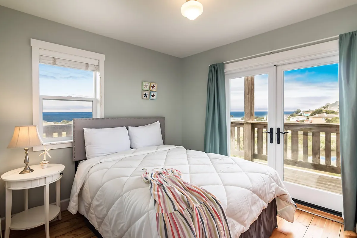 A peaceful bedroom with a bed dressed in white bedding, large windows that frame an expansive ocean view, and a nightstand with a lamp. The room is flooded with gentle morning light, creating a cozy nook perfect for relaxation or reading.