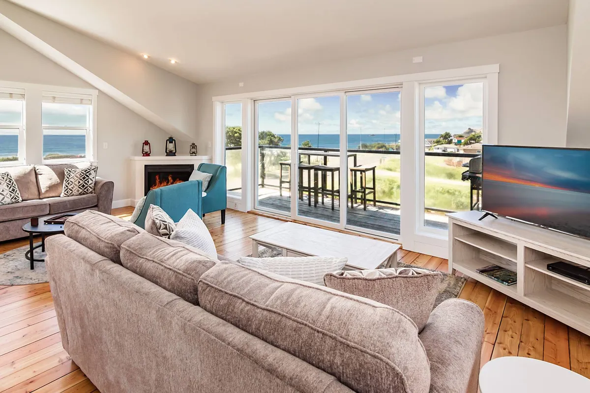 A welcoming great room with large windows framing sweeping ocean views, a comfortable seating area with plush sofas, a cozy fireplace, and a television. The scene is bathed in natural light, creating a warm and inviting space for relaxation, family gatherings, or game nights.