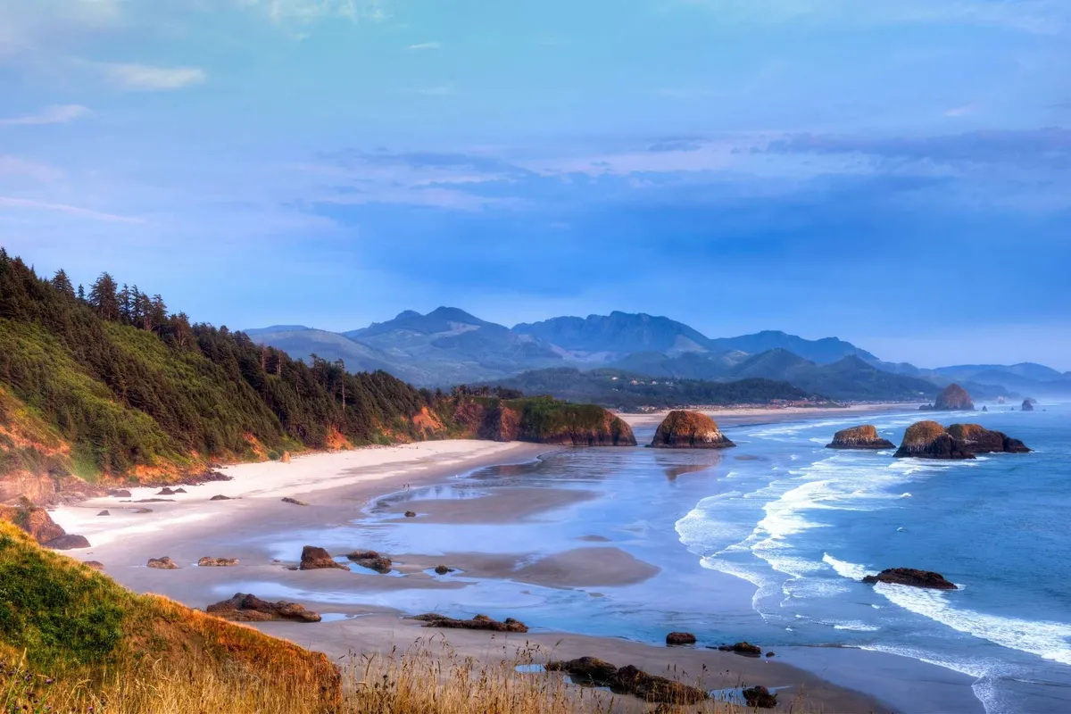 A sweeping view over rock formations, beach, ocean waves, and a distant mountain range. The scene is peaceful, with the sun setting over the water, and highlights the natural beauty that surrounds the property.