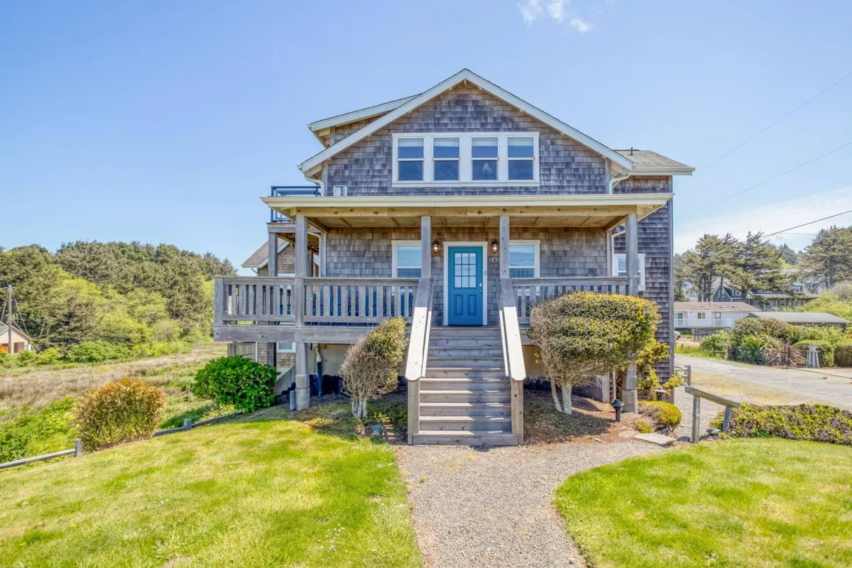 The image showcases the welcoming exterior of a beautifully updated single-family home, featuring a charming blue door, a spacious front porch with comfortable seating, and a carefully landscaped garden. The porch is perfectly positioned for relaxing, entertaining, and enjoying tranquil sunset views, while the lush surroundings evoke a serene coastal vibe.