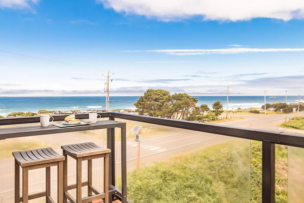 This image captures a tranquil moment on the balcony overlooking a lush wetland and the distant ocean. Two coffee cups and a snack plate rest on a table, suggesting a peaceful morning spent sipping coffee while taking in the expansive, nature-filled view. The balcony seating is positioned to maximize enjoyment of the panorama, with green vegetation and the open sky inviting wildlife viewing.