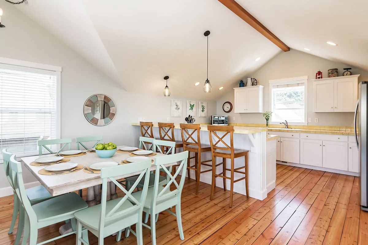 An inviting view of the dining area and kitchen, featuring a spacious dining table, kitchen island, bar stools, and stylish pendant lights. The layout suggests plenty of space for shared meals, board games, and lively conversation, with a seamless flow between cooking and dining areas.