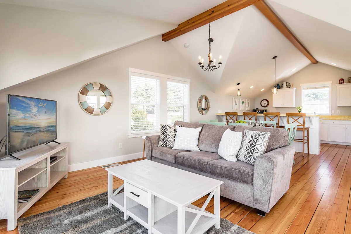 An inviting interior with a plush sofa oriented towards a large television, surrounded by a comfortable dining area and expansive windows. The space is softly lit by a chandelier, suggesting a multi-purpose room perfect for relaxing, watching TV, enjoying group games, or sharing meals.